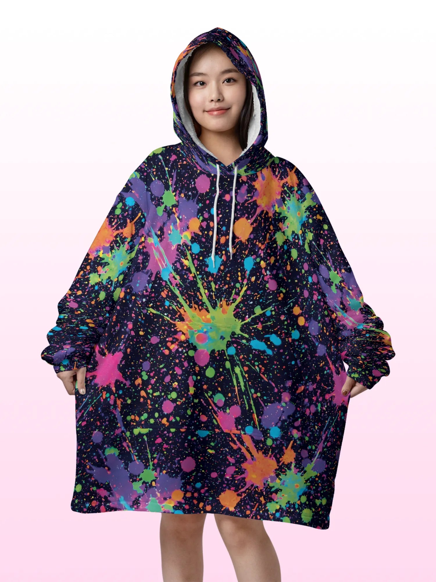 Front view of a woman wearing an oversized hooded blanket featuring a vibrant neon paint splatter design, large front kangaroo pocket, and plush white sherpa lining