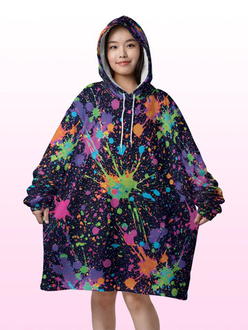 Front view of a woman wearing an oversized hooded blanket featuring a vibrant neon paint splatter design, large front kangaroo pocket, and plush white sherpa lining