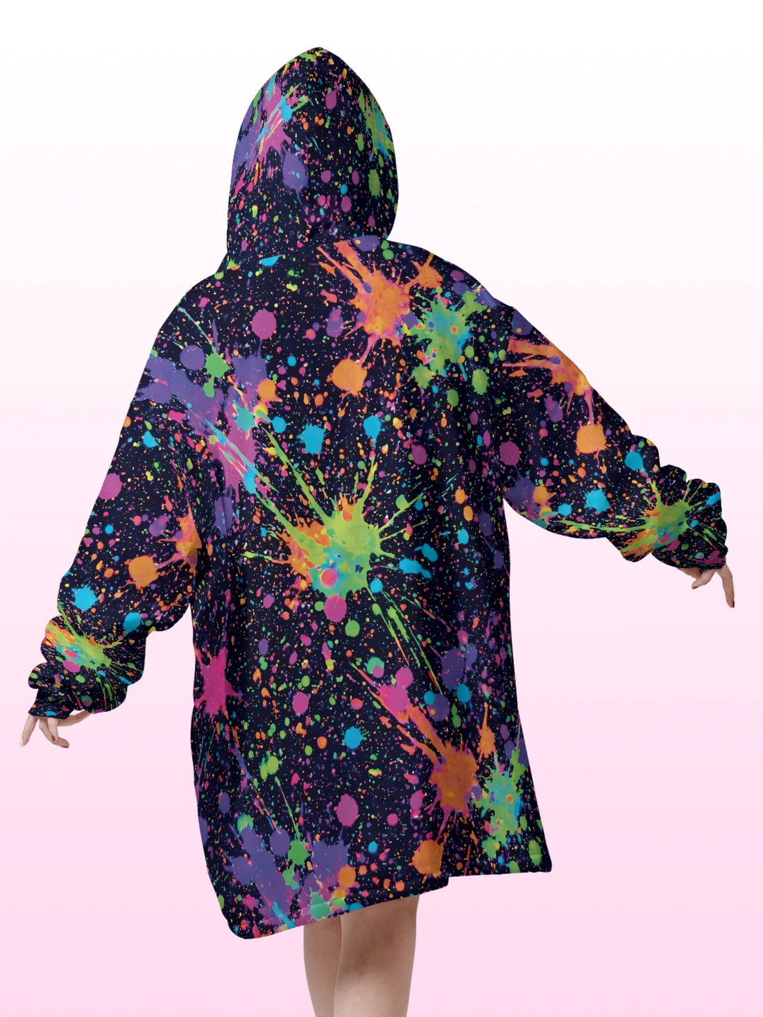 Front view of a woman wearing an oversized hooded blanket featuring a vibrant neon paint splatter design, large front kangaroo pocket, and plush white sherpa lining